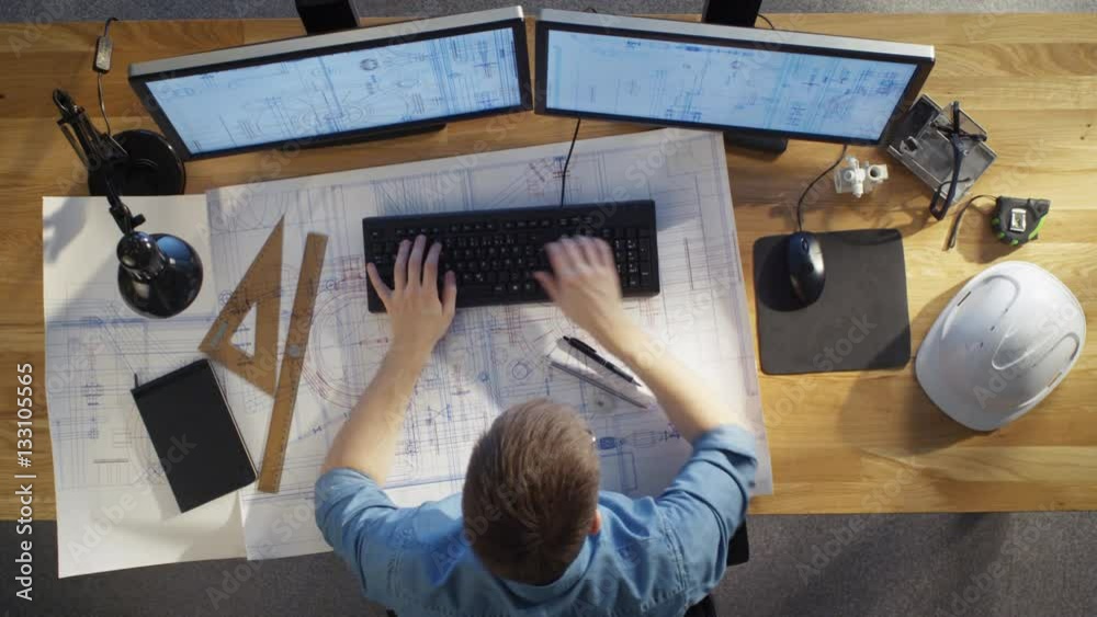 Top View of architectural Engineer Working on His Blueprints, Holding Tablet Computer, Using Desktop Computer Also. His Desk is Full of Useful Objects and Evening Sun. Top View of architectural Engineer Working on His Blueprints, Holding Tablet Computer, Using Desktop Computer Also. His Desk is Full of Useful Objects and Evening Sun.