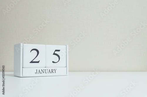 Closeup white wooden calendar with black 25 january word on blurred white wood desk and cream color wallpaper in room textured background with copy space , selective focus at the calendar