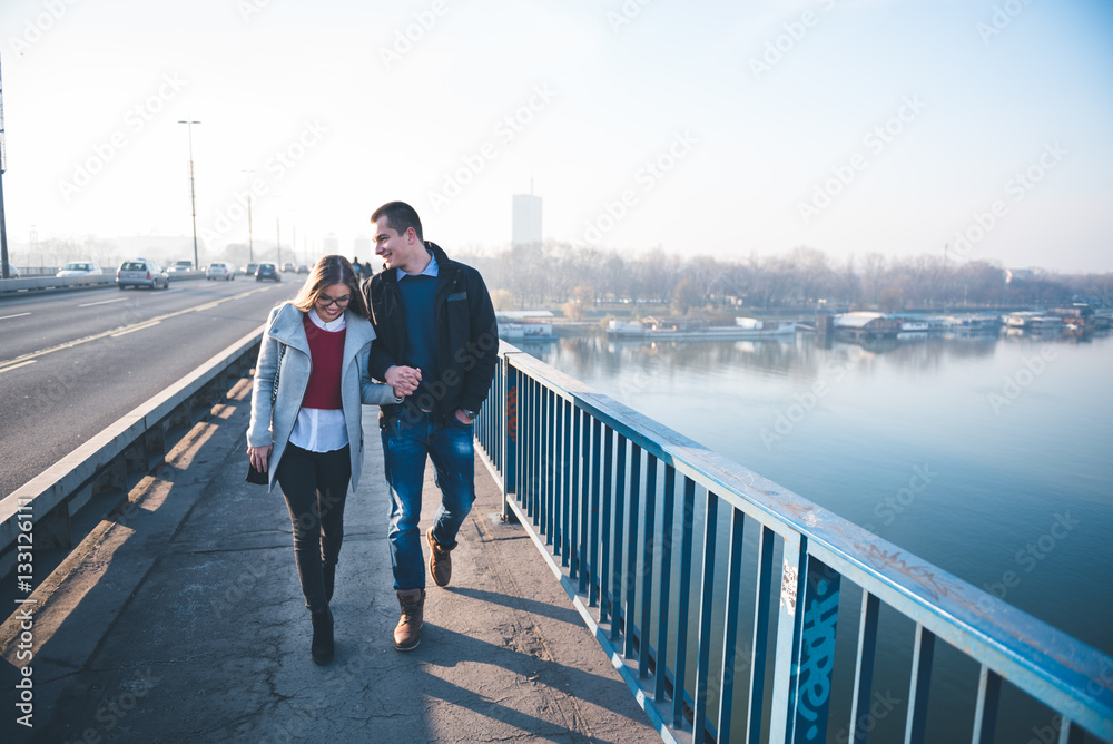 People in love theme. Attractive young couple enjoying in city outdoors. They walking on the city bridge, holding hands and smiling.