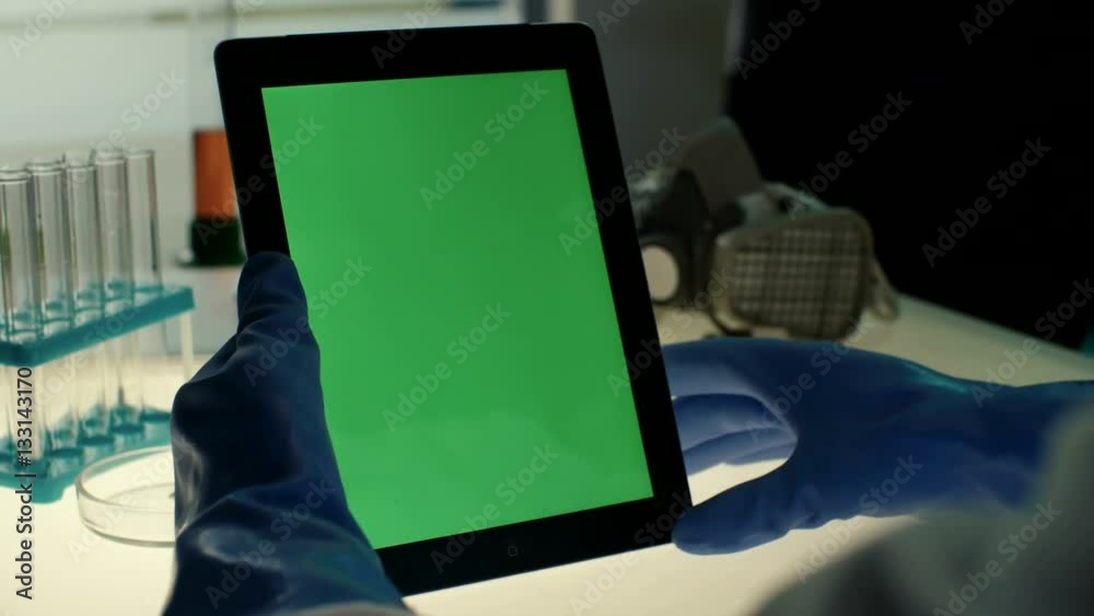 Scientist pressing in center of touch screen on tablet PC with green screen in modern laboratory on a background of test tubes and protective mask