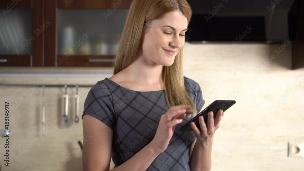 Beautiful young woman with a phablet standing in kitchen. Browsing Internet and smiling. Beautiful young woman with a phablet standing in kitchen. Browsing Internet and smiling.