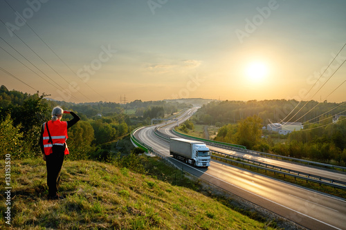 Obraz na płótnie The man in the orange safety vest observing the highway with a moving truck on a hill in the countryside at sunset. View from above. w salonie