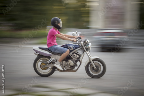 Man riding motorcycle. Young guy in a pink T-shirt and denim shorts, with a motorcycle helmet on his head quickly rides on a motorcycle on the street © OlegOk