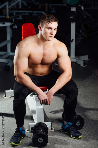 professional bodybuilder sitting on the bench, resting between exercises with dumbbells at gym. Big