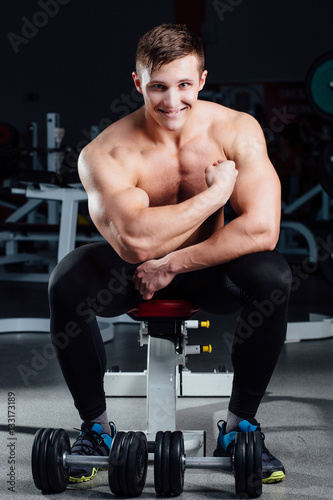 professional fitness bodybuilder sitting on the bench and demonstrates muscles of your body after a workout, at gym.