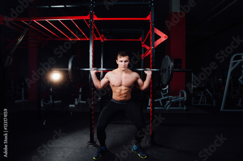 portrait of a handsome athlete at gym. man does the exercise with barbell, standing and squatting. big confident