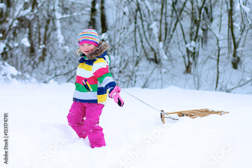 Portrait of beautiful toddler girl playing outdoors with snow. Happy little child wearing colorful knitted hat and blue coat enjoying winter day in the park or forest.