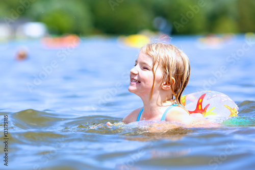 Happy little girl swimming in a lake using inflatable ring. Lovely kid splashing with water outdoors on hot summer day.