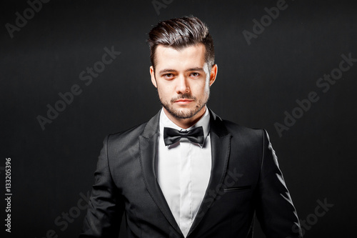 Fotografie, Obraz portrait of confident handsome man in black suit with bowtie posing in dark stud