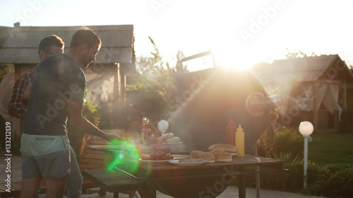 Two Caucasian men putting fried sausages on grill grid with beer in hands. In slowmotion
