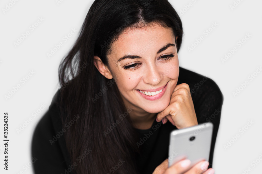 Close up portrait of attractive young happy woman with smart phone with healthy toothy smile,with hand on chin, reading or text message, on light grey background. Technology and business theme. Close up portrait of attractive young happy woman with smart phone with healthy toothy smile,with hand on chin, reading or text message, on light grey background. Technology and business theme.