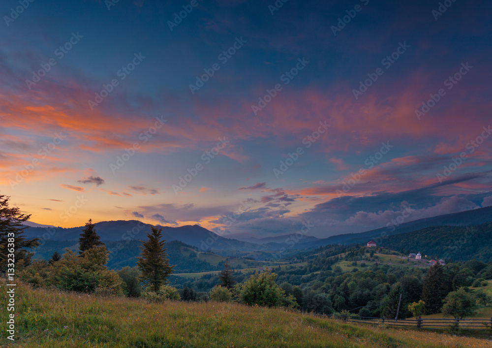Beautiful summer sunset landscape in Carpathian mountains. Ukraine. Green pasture with wildflowers and shepherds house in the middle ground. Small private hotel as small detail far away.