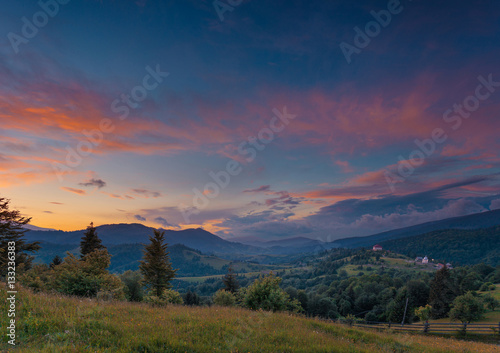 Beautiful summer sunset landscape in Carpathian mountains. Ukraine. Green pasture with wildflowers and shepherds house in the middle ground. Small private hotel as small detail far away. © stone36