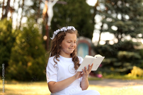 Pierwsza komunia, dziewczynka w białej sukience z książeczką na tle kapliczki.