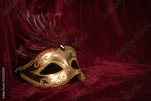 Golden venetian carnival mask with red feathers seen from the side on a draped red velvet theater curtain 