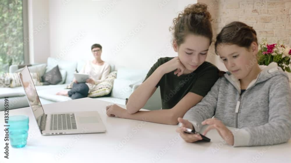Sisters using smartphone together with Mom in background Sisters using smartphone together with Mom in background