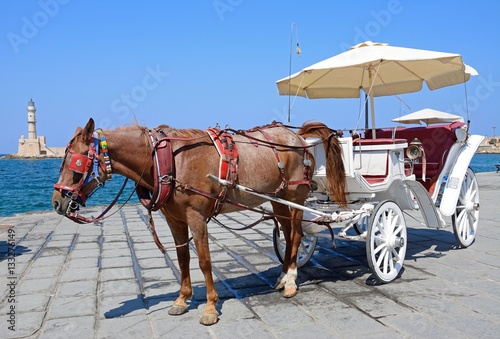 Horse drawn carriage on the quayside with the Venetian lighthouse at the harbour entrance to the rear, Chania, Crete. © arenaphotouk