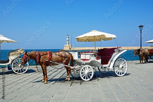 Horse drawn carriages on the quayside with the Venetian lighthouse at the harbour entrance to the rear, Chania, Crete.