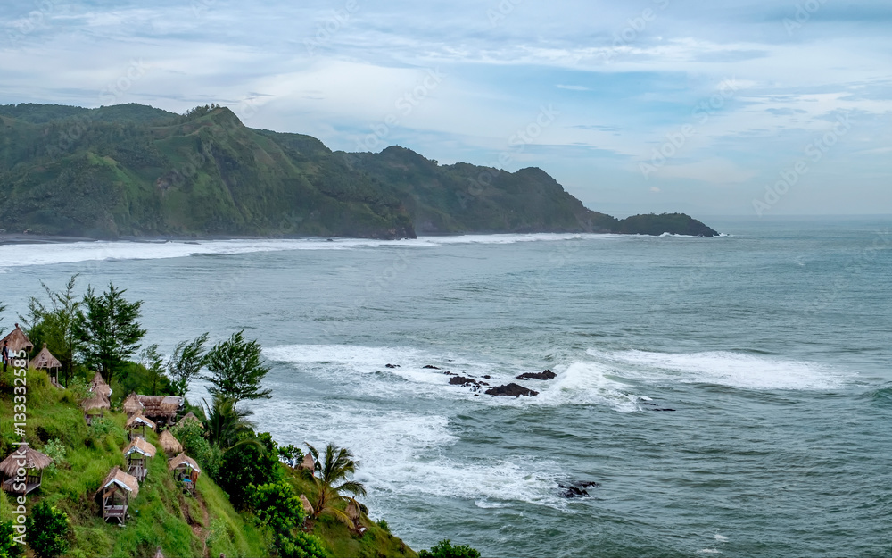 Photo of sea and land from top of hill, with little hut or gazebo on the side of the hill. And waves hit the shore in the bottom. Captured on Menganti Beach, Kebumen, Indonesia