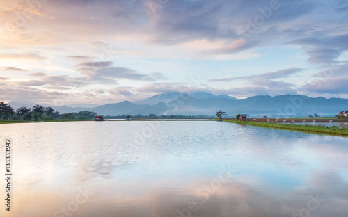 Photo of very vast, broad, large, spacious pond, stretched into the horizon. Behind it is a line of hills and mountains that also expansive, and beautiful cloud purple sky. This photo captured at sunr