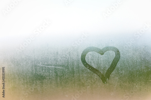 Abstract blurred love heart symbol drawn by hand on the wet frozen window glass with sunlight background. photo