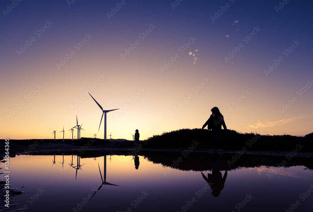 Wind turbines at a wind farm with the silhouettes of two girls reflected on the water surface at sunset, Crete, Greece