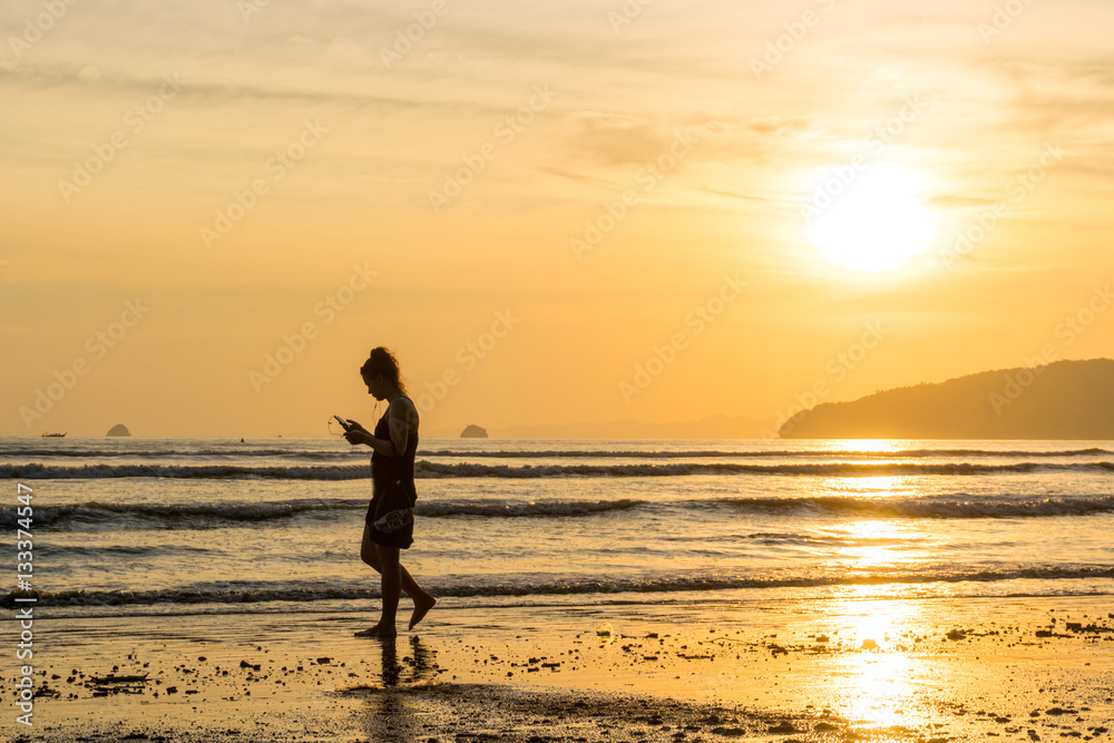 Young woman walking with smartphone on the beach against the backdrop of a beautiful sunset
