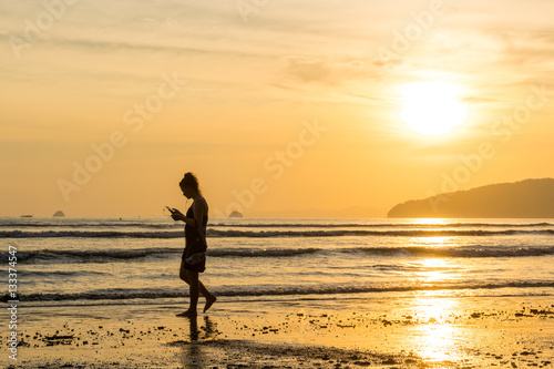 Young woman walking with smartphone on the beach against the backdrop of a beautiful sunset