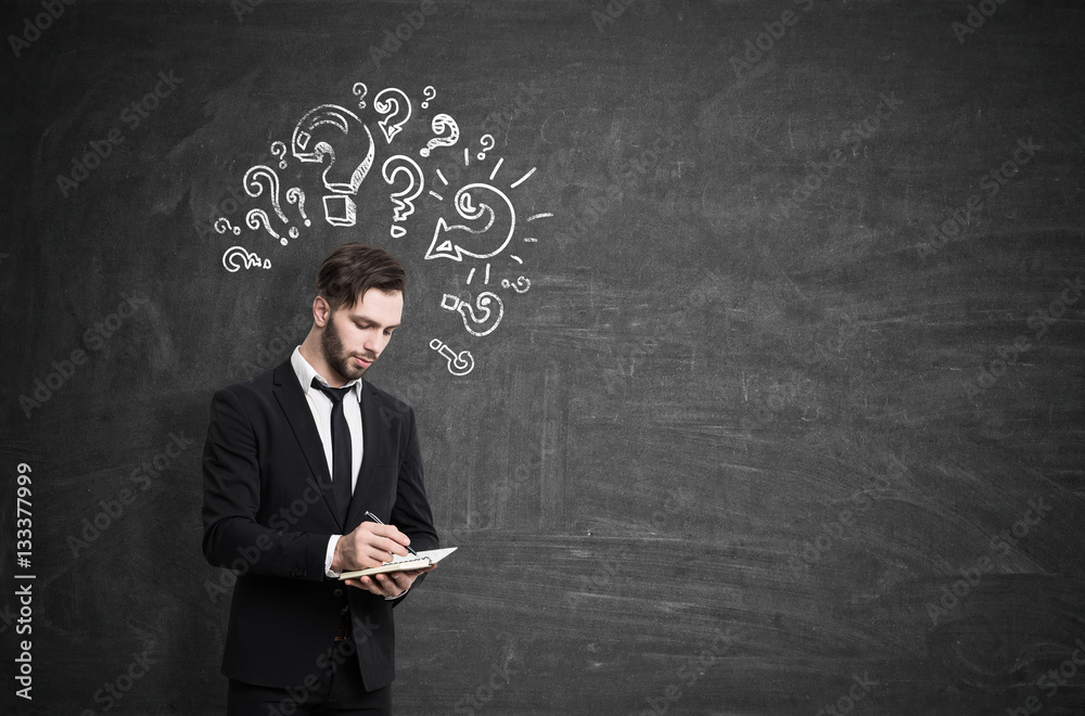 Portrait of a thoughtful businessman in a black suit standing near a chalkboard with question marks drawing on it. Mock up Portrait of a thoughtful businessman in a black suit standing near a chalkboard with question marks drawing on it. Mock up