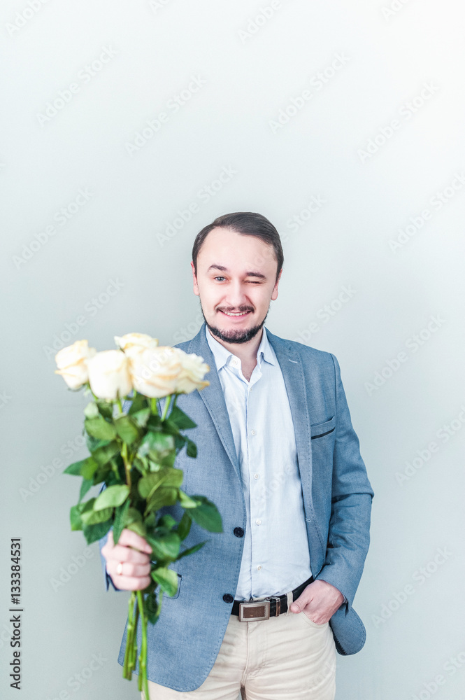 Handsome man with beard standing against a grey background with a bouquet of white roses. Business man gives roses to the occasion Handsome man with beard standing against a grey background with a bouquet of white roses. Business man gives roses to the occasion