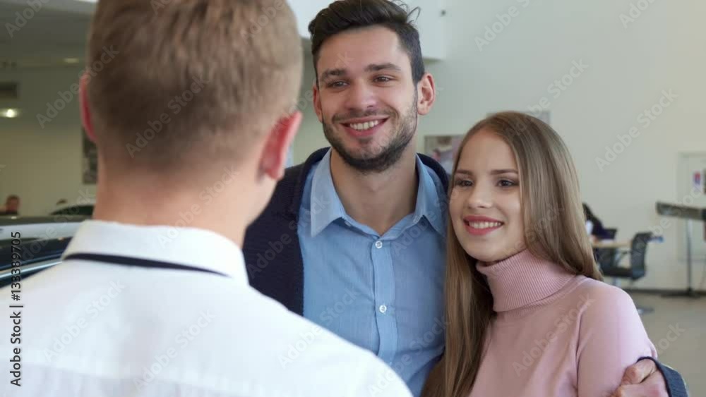 Pretty caucasian woman getting the car key from the salesman at the dealership. Handsome brunette man embracing his attractive girlfriend. Young brunette bearded man and his brown haired girlfriend