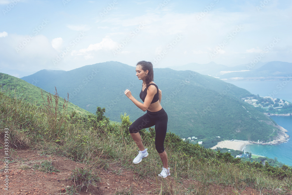 Fit female jogger exercising, running uphill with sea and mountains in background Fit female jogger exercising, running uphill with sea and mountains in background