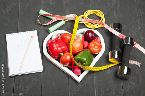 Sport and diet. Healthy lifestyle. Vegetables  dumbbells  notebook. Peppers  tomatoes  garlic  onion  radish in a heart on  black background