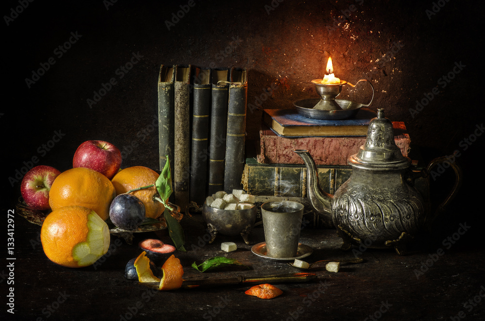 Obraz premium Classic still life with vintage silver plate full of oranges,apples and plums placed with elegant old kettle,sugar,cup of tea,old books and illuminated candle..