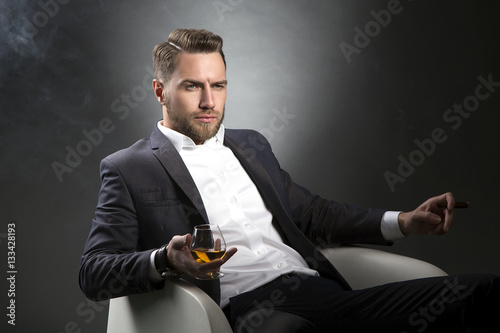 Portrait of young man with pefect hair style drinking cognac and smoking cigar photo