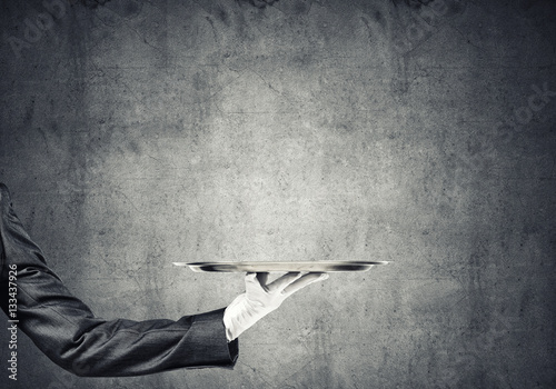 Hand of butler holding empty metal tray against concrete background photo