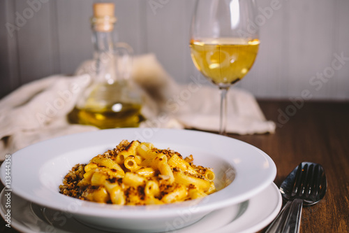 Cheese flavored rigatoni pasta in a white plate on a wooden table with kitchen appliances, glass of dry white wine and glass jug with olive oil on the old gray kitchen towel in the background