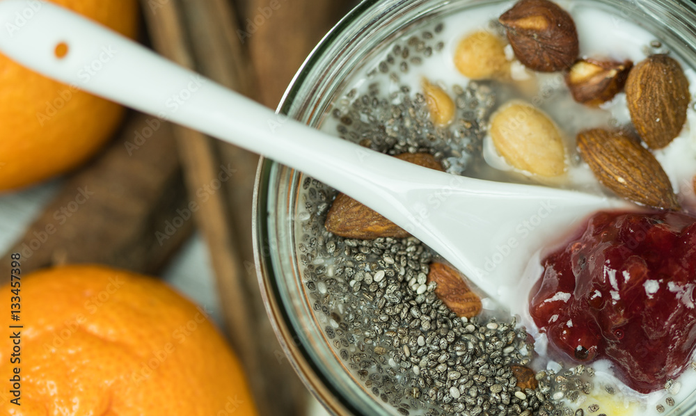 Fototapeta premium Macro shot of a white ceramic teaspoon of strawberry jam dipped in healthy breakfast mix, almonds, hazelnuts and walnuts chia seeds and cereal. Tangerines and Cinnamon sticks in the background.