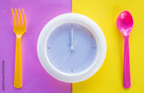white clock on coloured background  at dinner spoon and fork waiting for food