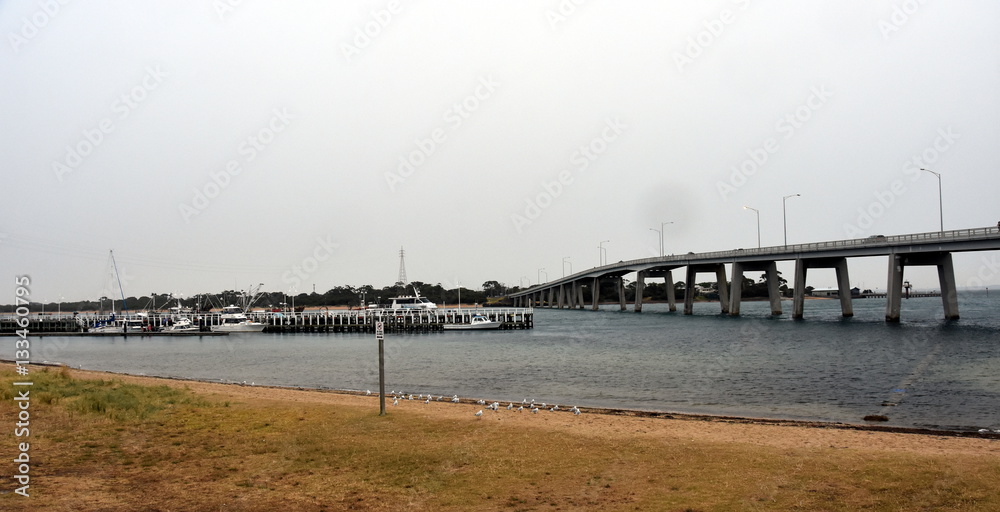 Panorama at the concrete bridge to Phillip Island from San Remo in Victoria, Australia with white fishing boats at jetty, green grass and green bush on shore of Phillip Island. Panorama at the concrete bridge to Phillip Island from San Remo in Victoria, Australia with white fishing boats at jetty, green grass and green bush on shore of Phillip Island.