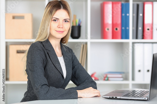 Businesswoman sitting at a desk in the office right shoulder forward. Hand on the keyboard. Looking into the camera.