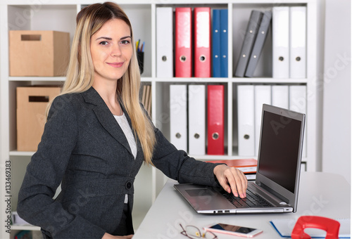 Businesswoman sitting at a desk in the office right shoulder forward. Hand on the keyboard. Looking into the camera.