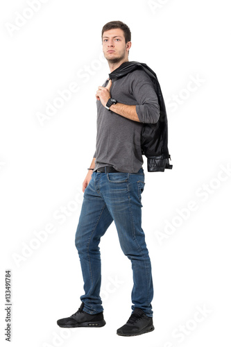 Side view of casual young man carrying jacket over shoulder looking at camera. Full body length portrait isolated over white background.