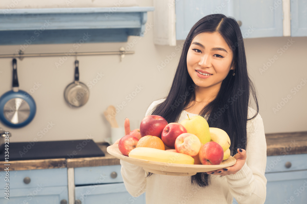 Toned picture of Chinese or Korean lady posing with bowl full of different fruits. Beautiful woman smiling for camera and showing fruits to camera at home. Toned picture of Chinese or Korean lady posing with bowl full of different fruits. Beautiful woman smiling for camera and showing fruits to camera at home.