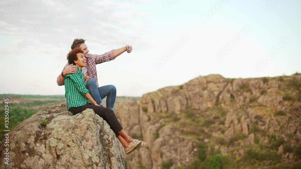 Interracial couple sitting on big rock making selfie with mobile phone smiling posing in slowmotion Interracial couple sitting on big rock making selfie with mobile phone smiling posing in slowmotion