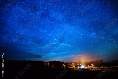 Camping fire under the amazing blue starry sky with a lot of shining stars and clouds. Travel recreational outdoor activity concept.
