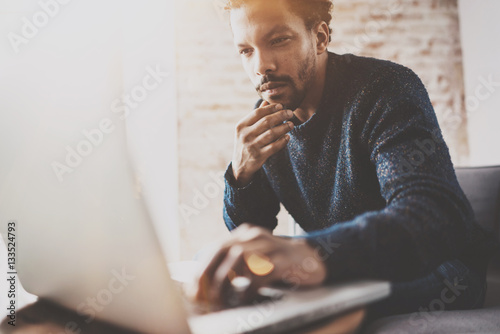 Pensive young African businessman using laptop while sitting on sofa at his modern coworking place.Concept of business people.Blurred background,color filter,flare effect.