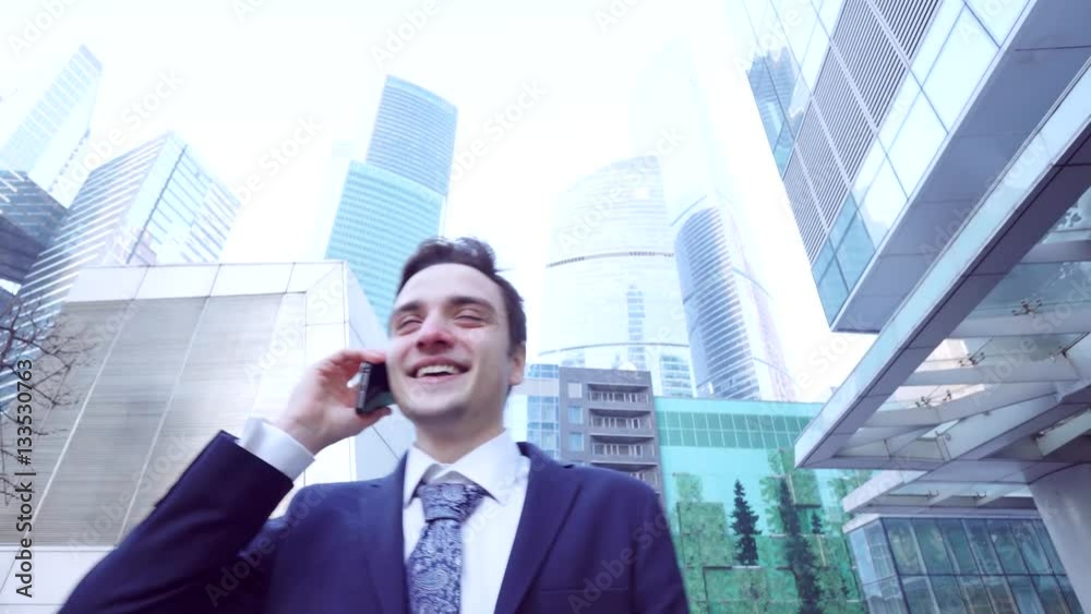 Casual young business talking on smart phone at day. Handsome young business man talking on smartphone and walking towards camera smiling happy wearing suit jacket outdoors
