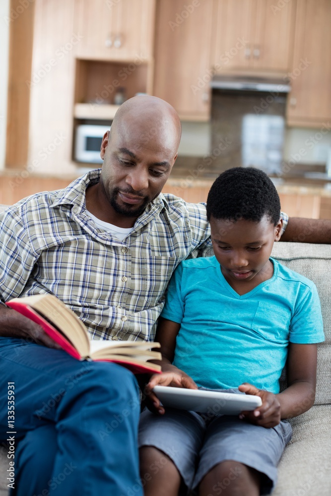 Father reading book while son sitting next to him using digital 