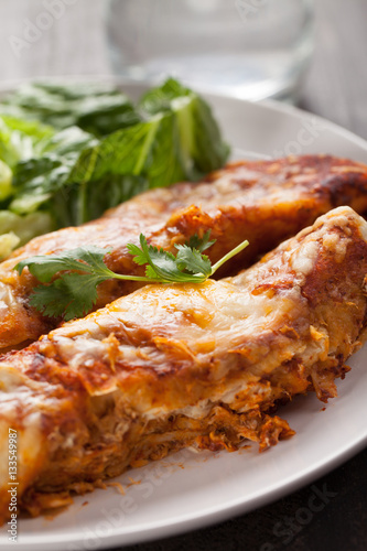 Spicy chicken enchiladas with a side of light green salad on a dark wood background fresh from oven
